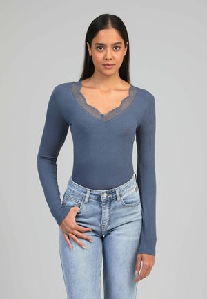 Femme portant un haut bleu ajusté à manches longues avec un détail en dentelle au col en V et un jean taille haute bleu clair, posant devant un fond gris uni.