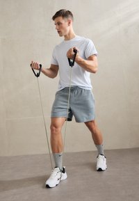 Benzile de rezistență cu mânere negre și elastic verde, utilizate de un bărbat îmbrăcat într-un tricou gri deschis și șorturi, care execută un exercițiu.