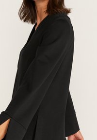 Černý dlouhý rukáv s výstřihem do V, s volněj ším střihem, širokými manžetami a hladkou strukturou. Viditelný postranní rozparek.