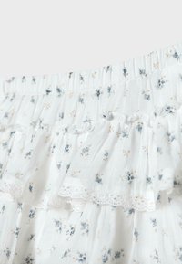 Tissu blanc avec petit imprimé floral bleu et beige, taille élastiquée froncée et détail de bordure en dentelle à volants.