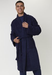 Junger Mann, der lächelt und einen dunkelblauen Bademantel trägt, der an der Taille gebunden ist, während er mit einer Hand den Kragen anpasst.