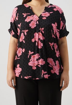 Femme portant un chemisier noir à manches courtes retroussées avec un imprimé floral rose, associé à un pantalon noir, sur un fond uni.