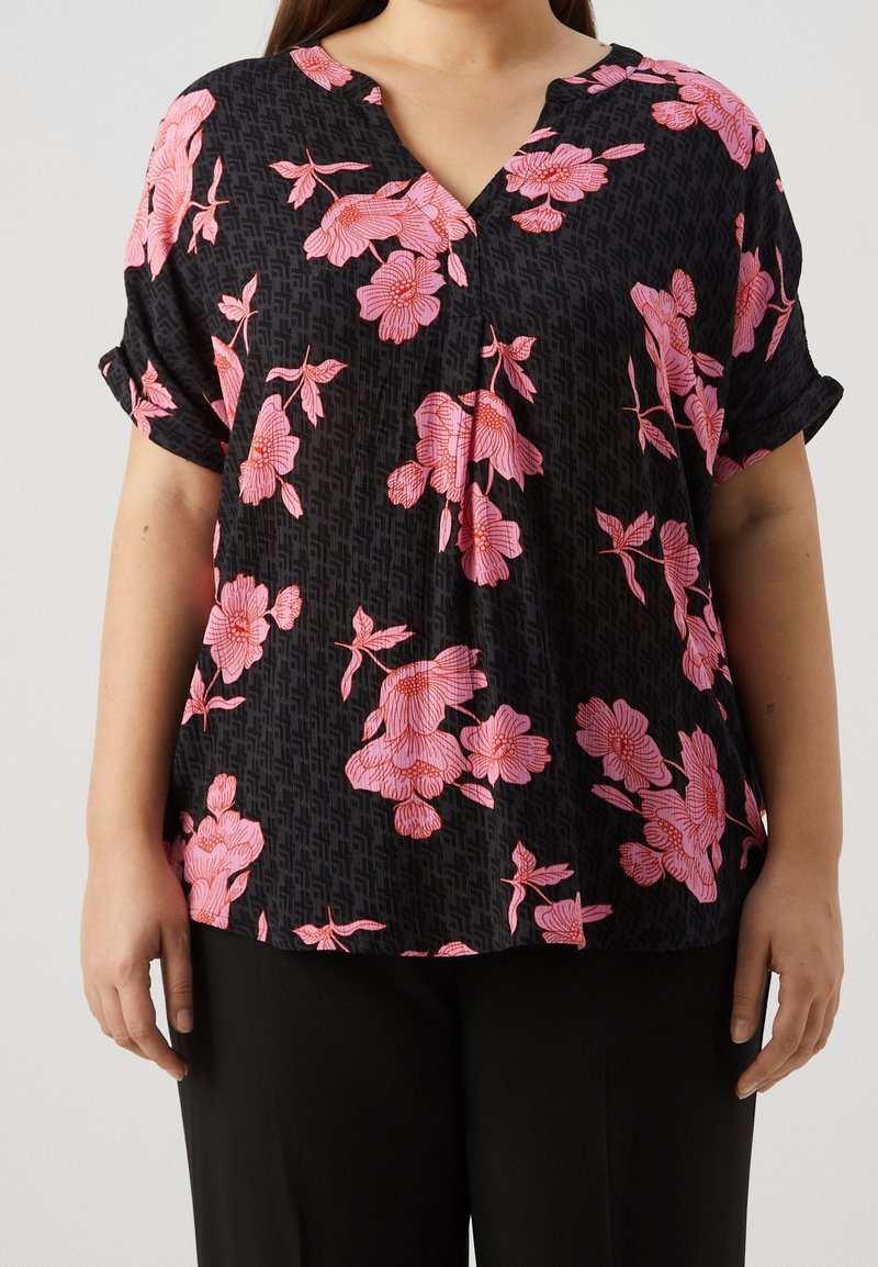 Femme portant un chemisier noir à manches courtes retroussées avec un imprimé floral rose, associé à un pantalon noir, sur un fond uni.