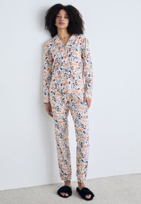 Blumiges Pyjama-Set bestehend aus einem Hemd mit Knöpfen und einer locker sitzenden Hose, aus weichem Stoff mit einem Orange-, Blau- und Weißdruck. Schwarze flauschige Hausschuhe.
