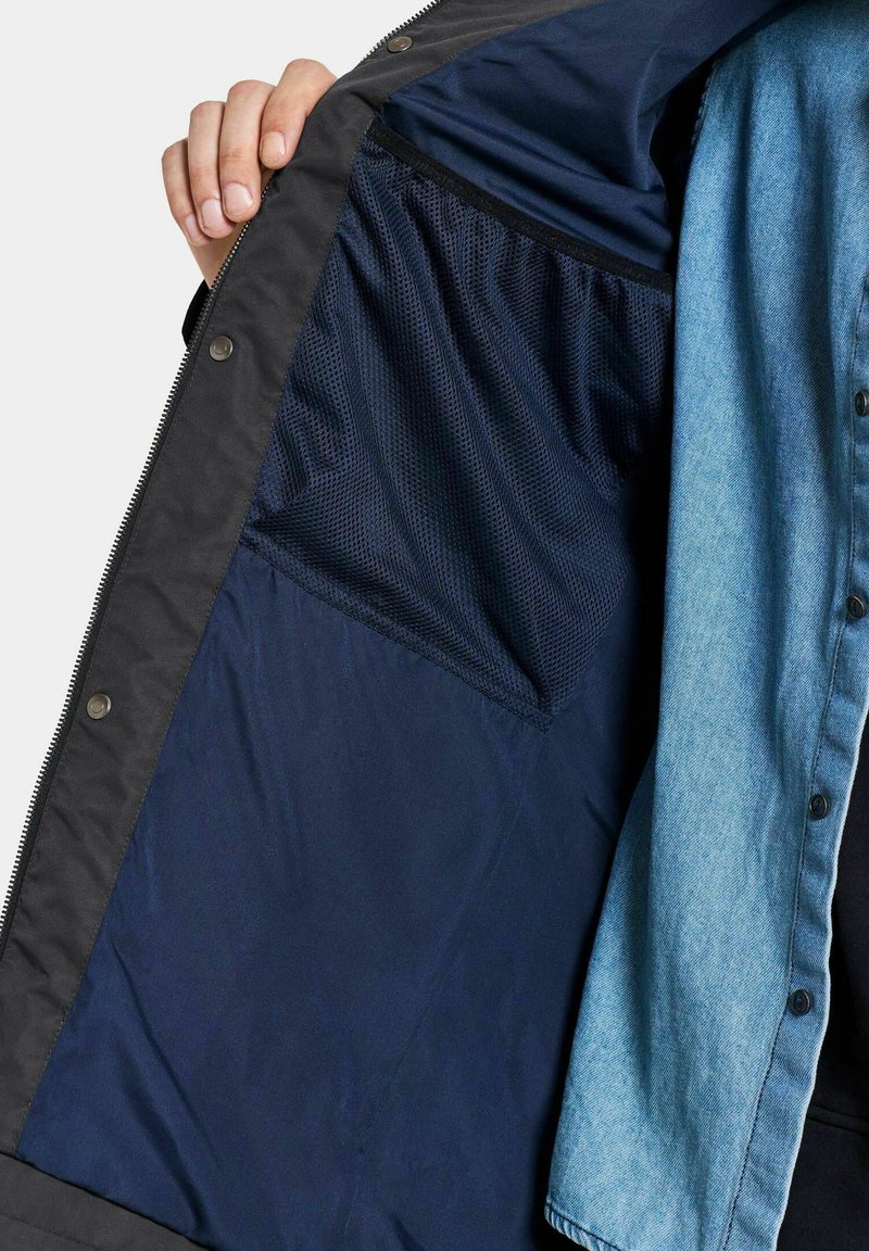 Hand, die eine offene schwarze Jacke halten, die eine marineblaue Netzinnentasche und einen blauen Jeanshemdärmel darunter zeigt.