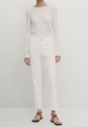 Woman wearing a white long-sleeve top, white slim-fit pants, and black flat sandals with brown ankle straps against a plain background.