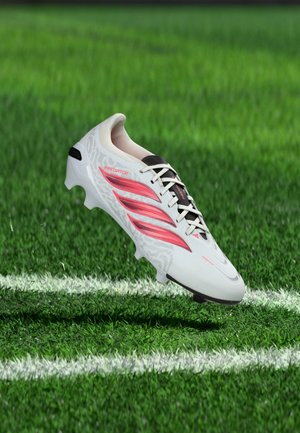 Weißer Fußballschuh mit roten Streifen beschriftet mit "Predator", schwebt über grünem Gras in der Nähe weißer Spielfeldlinien.