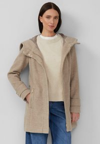 Manteau beige à capuche avec une texture douce, présentant des accents de quincaillerie dorée, des poches zippées et une coupe ample, porté par-dessus un pull léger et un jean.