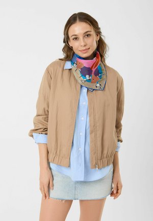 Femme portant une veste beige sur une chemise bleu clair et une jupe en jean avec une écharpe colorée à motifs autour du cou, posant sur un fond uni.