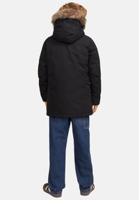 Svart parkas med en foderhuva av päls, kviltad design och manschetter. Åtföljs av löst sittande blå denimjeans. Bakifrån.