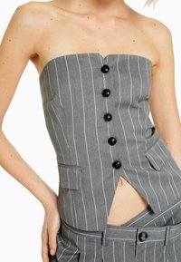 Kobieta w szarej, paskowanej bezrękawniczce z czarnymi guzikami oraz pasujących spodniach w paski, z ręką opartą na biodrze, odsłoniętym brzuchem.