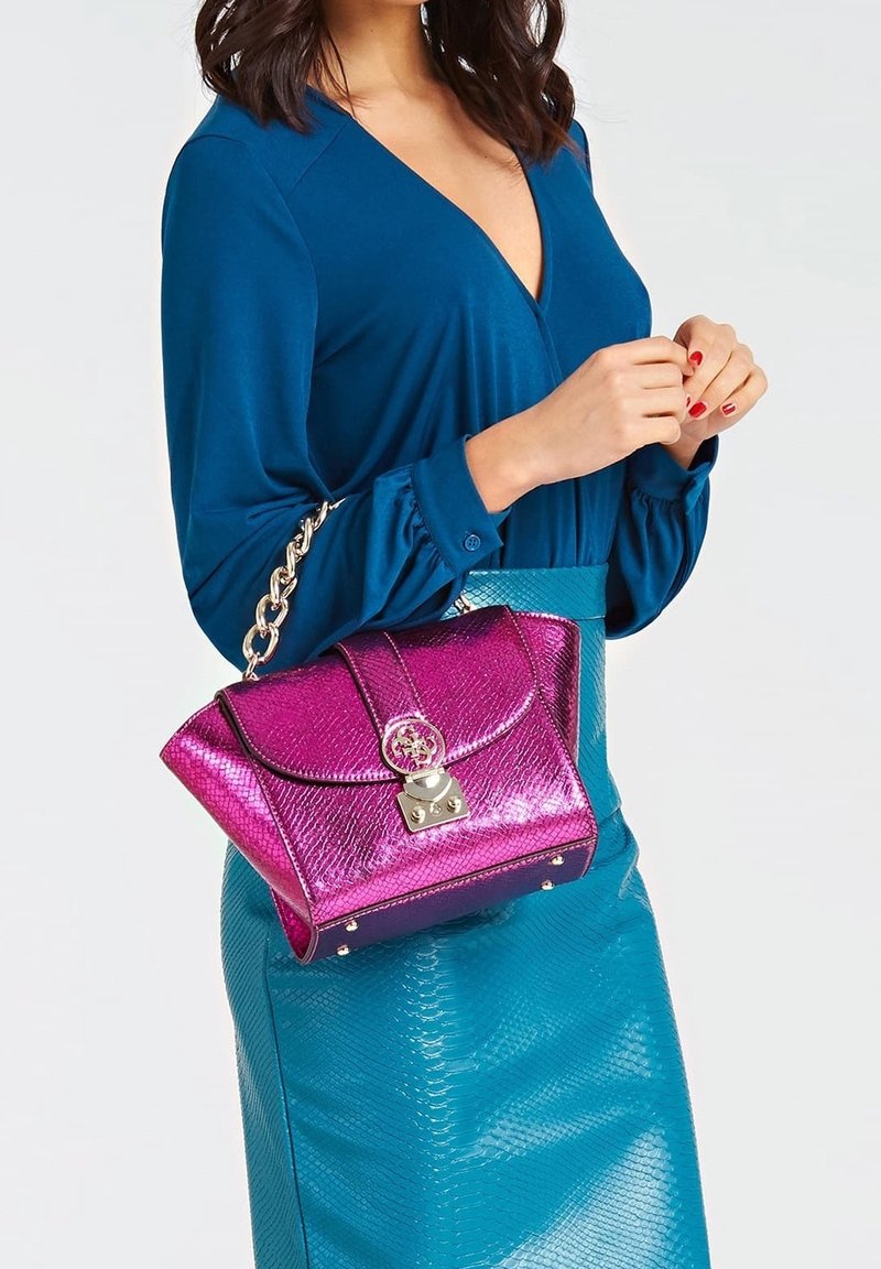 Femme portant une robe bleue tient un sac à main rose brillant avec une chaîne et un fermoir dorés, montrant le haut du corps et les mains croisées.