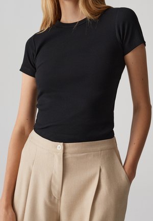 Femme portant un haut noir côtelé à manches courtes ajusté, rentré dans un pantalon beige plissé taille haute avec une fermeture par bouton.