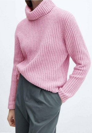 Vrouw draagt een roze geribbelde coltrui die in een high-waisted grijze broek is gestopt, met één hand in de zak.