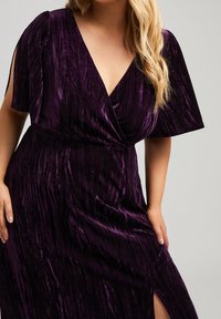 Lila, strukturerad sammetklänning med djup V-ringning, korta volangärmar och slits på sidan, som visar upp en flödande design och mjuk lyster.