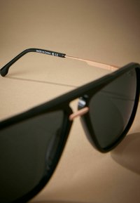 Schwarze Sonnenbrille mit dunklen Gläsern, die ein mattes Finish und ein schlankes Design aufweisen. Sie haben einen goldenen Akzent am Scharnier und ein strukturiertes schwarzes Bügel.