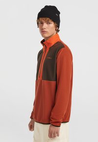 Fleece-Pullover in Orange und Braun mit einem halben Reißverschlusskragen, langen Ärmeln und einer strukturierten Oberfläche. Getragen mit einer schwarzen Mütze, mit minimal sichtbarem Branding.