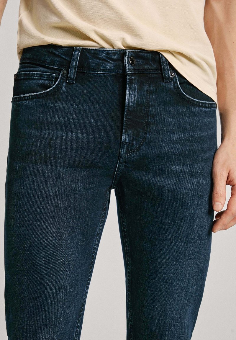 Jeans de mezclilla azul oscuro con un diseño ajustado, que cuentan con cinco bolsillos, pasadores para cinturón y un cierre de botón. Textura suave, con un desvanecimiento mínimo.