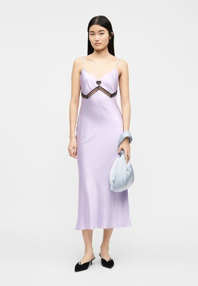 Γυναίκα με φόρεμα slip από σατέν λεβάντας με μαύρη δαντέλα, κρατώντας τσαντάκι σε ανοιχτό μπλε χρώμα, φορώντας μαύρες μυτερές γόβες με χαμηλό τακούνι, στέκεται σε λευκό φόντο.