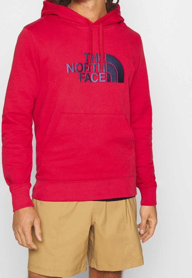 Homme portant un sweat à capuche The North Face rouge vif et un short beige, debout devant un fond clair uni.