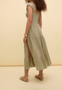 Robe longue de couleur vert clair avec un motif texturé, bretelles froncées et une jupe à étages avec une fente sur la cuisse. Associée à des sandales dorées.