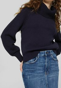 Pull en laine bleu marine avec un col texturé haut et des poignets côtelés. Présente un corps lisse et des fentes latérales, associé à une jupe en denim bleu.