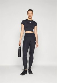 Jonge vrouw in zwarte sportkleding houdt een zwarte waterfles vast, staande tegen een lichte achtergrond.