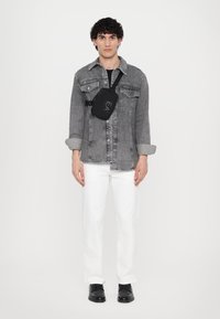 Black crossbody bag with logo, worn over a gray denim jacket, white pants, and black shoes. Model stands against a plain background.