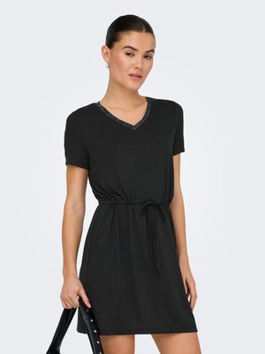 Femme portant une robe noire à manches courtes avec un col en V et une taille à cordon, tenant un sac à main noir sur un fond clair.