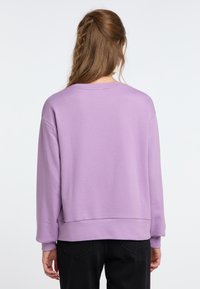 Felpa di cotone viola con collo a giro, spalle scese e polsini a costine. Tessuto liscio e vestibilità rilassata, abbinata a pantaloni neri.