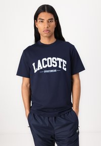 Navy zila kokvilnas T-krekls ar baltu "LACOSTE" logo un "SPORTSWEAR" zem tā. Apaļš dekoltē un īsas piedurknes. Apvienots ar tumšiem biksēm.