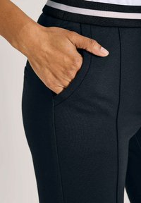Hand met een gemiddelde huidtint gestoken in de zijzak van zwarte, slim fit broek met een tailleband met witte en zwarte strepen.