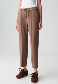 Pantalons marron sur mesure avec des plis sur le devant et une taille à cordon, portés avec des baskets marron foncé et un haut sans manches blanc.