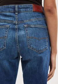 Tamsiai mėlynos spalvos aukštos juosmens džinsai iš džinso audinio, su rudos odos prekės ženklo lipduku, dviem užpakalinėmis kišenėmis ir kontrastingu oranžiniu siuvimu.