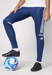 Jūras spēku sporta legingi ar baltām sānu svītrām, gluda tekstūra, pieguļošs dizains, kopā ar melniem sporta apaviem uz rakstainas futbola bumbas.