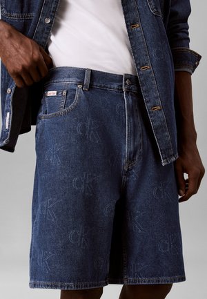 Homme portant un short en denim bleu foncé avec des logos "cK" subtils, une chemise blanche et une veste en denim bleu foncé assortie, ajustant son short.