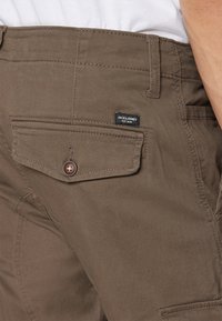 Pantalones de algodón marrones con un bolsillo trasero con botón, detalles de costura y una pequeña etiqueta de marca en la cinturilla.