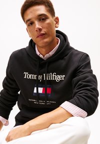 Felpa nera con cappuccio con logo "Tommy Hilfiger" e design della bandiera in rosso, bianco e blu. Indossata sopra una maglia a righe bianche e rosse.