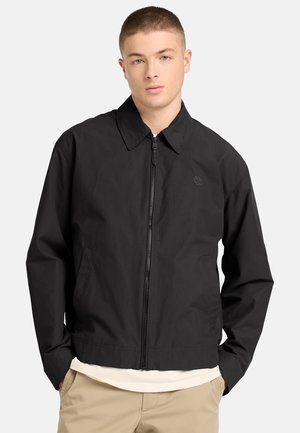 Ung mand iført en sort lynlåsjakke med krave og beige bukser, hænderne i lommerne, foran en ensfarvet lys baggrund.