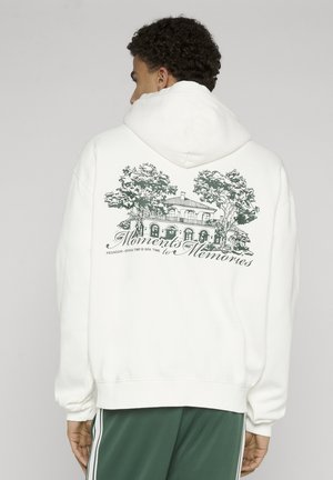 Person iført hvid hættetrøje med sort skitse af hus og træer samt teksten "Moments to Memories" på ryggen, set bagfra.