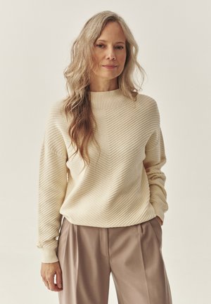 Beige ribbet sweater med høj hals og lange ærmer, parret med lysebrune plisserede bukser, sat mod en neutral baggrund.