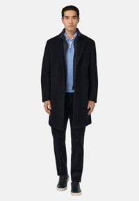 Tumedast villasegust mantel, millel on üks rind, sobitatud helesinise sviitriga ja tumedatest korduroodidest pükstega. Mustad spordijalanõud täiendavad välimust.