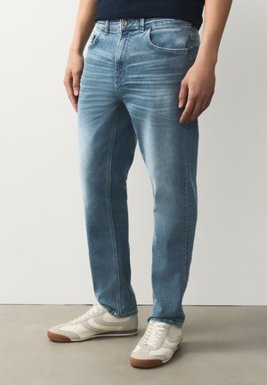 Uomo con jeans azzurri a gamba dritta e sneakers bianche, in piedi su un pavimento grigio contro una parete bianca.