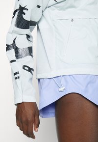 Personne portant une veste blanche avec un imprimé graphique noir sur les manches et un short violet clair, montrant une poche avec fermeture éclair et un cordon de serrage réglable.