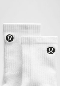 Weiße Sportsocken mit geripptem Muster, versehen mit einem aufgenähten schwarzen runden Logo am Bündchen. Einfaches, funktionales Design.