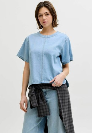 Mladá žena v světle modrém džínovém topu s krátkými rukávy, světle modrých džínách a černé kostkované košili uvázané kolem pasu.