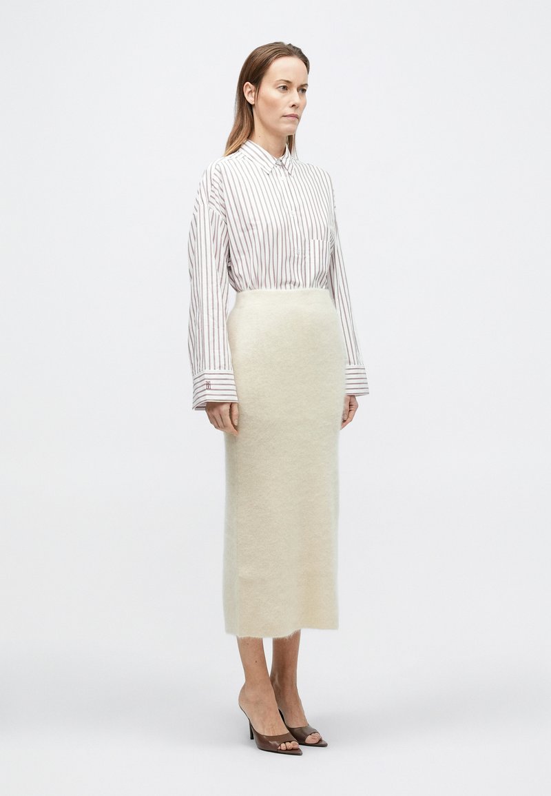 Woman standing wearing a white and red striped long-sleeve shirt, cream-colored midi skirt, and brown open-toe heels on a plain background.