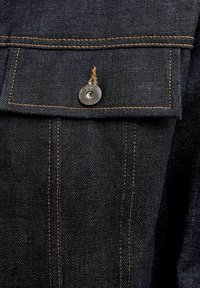 Nærbilde av mørk denimlomme med oransje søm og en metallknapp inngravert med "Jack & Jones Authentic Jeanswear."