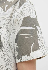 Cămașă cu mânecă scurtă, realizată dintr-un material ușor, cu un fundal alb și modele mari de frunze tropicale gri, prezentând o țesătură texturată.