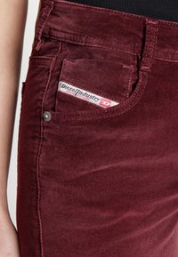 Pantalones de pana burdeos con un bolsillo frontal que presenta una etiqueta visible de Diesel. Los detalles de costura a lo largo de las costuras son prominentes, añadiendo textura.